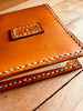 Close up of angled view of a closed handcrafted tan leather notepad cover on a wooden surface, featuring a personalized initial tab on the front and prominent white stitching