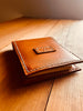 Angled view of a closed handcrafted tan leather notepad cover on a wooden surface, featuring a personalized initial tab on the front and prominent white stitching
