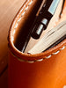 Close-up of the interior of a handcrafted tan leather notepad cover, showcasing the detailed white hand-stitching along the edge and a black pen secured in the pen holder next to a notepad.