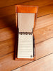 Full view of open handcrafted tan leather 3x5 notepad cover on a wooden surface, revealing a lined notepad on the right and a pen in its holder.