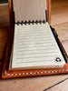 Open view of a handcrafted tan leather 3x5 notepad cover on a wooden surface, revealing a lined notepad on the right and a pen in its holder.