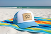 Light blue and white trucker hat with 'LIVE SALTY & STAY BREEZY' burlap patch resting on a colorful striped beach towel on sunny white sand with ocean waves in the background.